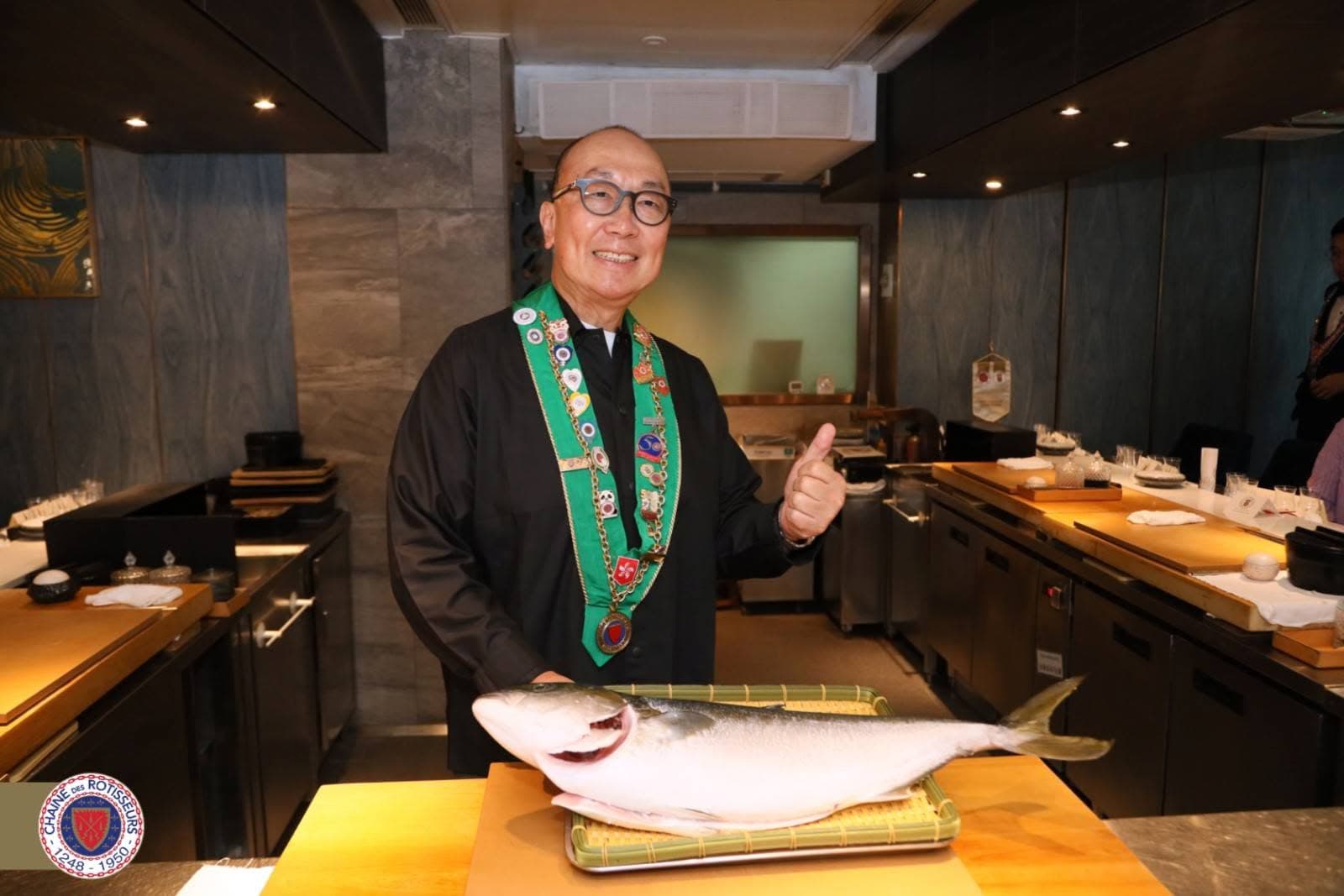 Jacky Kam in Chaîne des Rôtisseurs regalia giving a thumbs up in the kitchen